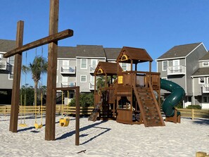 A wooden playground with a slide and swings, set against a backdrop of residential buildings.