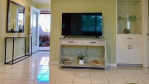 A living room with a TV on a white console, a glass cabinet, and a door leading to a balcony. - Diamond Head/Waikiki Surf Shack 2-Bedroom Private Ohana Unit (Honolulu)