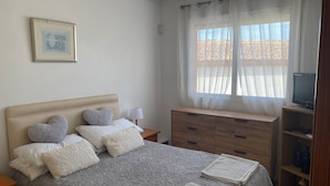 Un dormitorio con cama, mesita de noche, cómoda, televisor y ventana con cortinas ligeras.