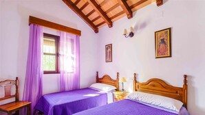 A room with two beds, wooden headboards, a window with purple curtains, a wooden chair, and a wall-mounted lamp. - Villas Atlantico Sur (Conil de la Frontera)