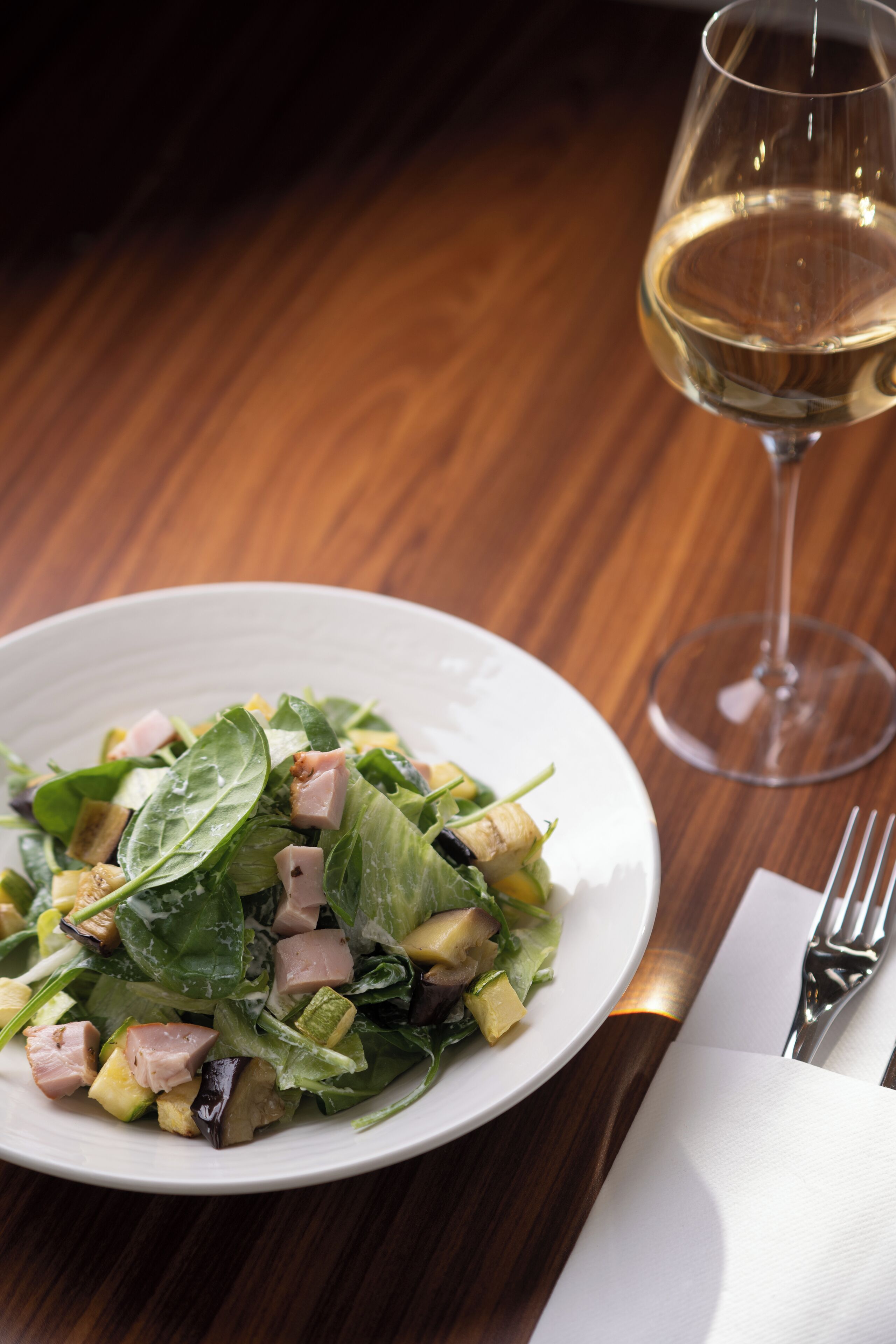 A white plate with a salad, a glass of white wine, and a fork on a wooden table.