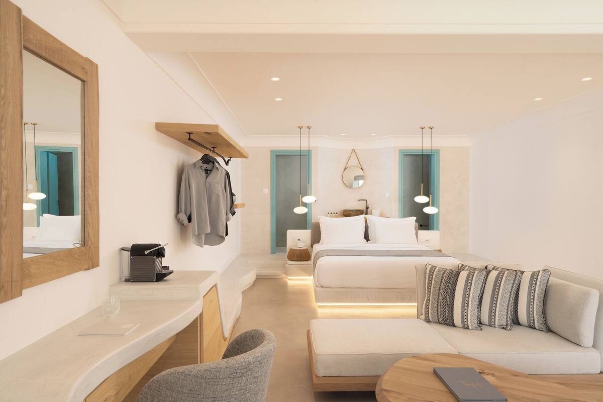 A modern hotel room with a large mirror, a sofa, a bed with striped pillows, a wooden shelf, and a bedside table with a glass.