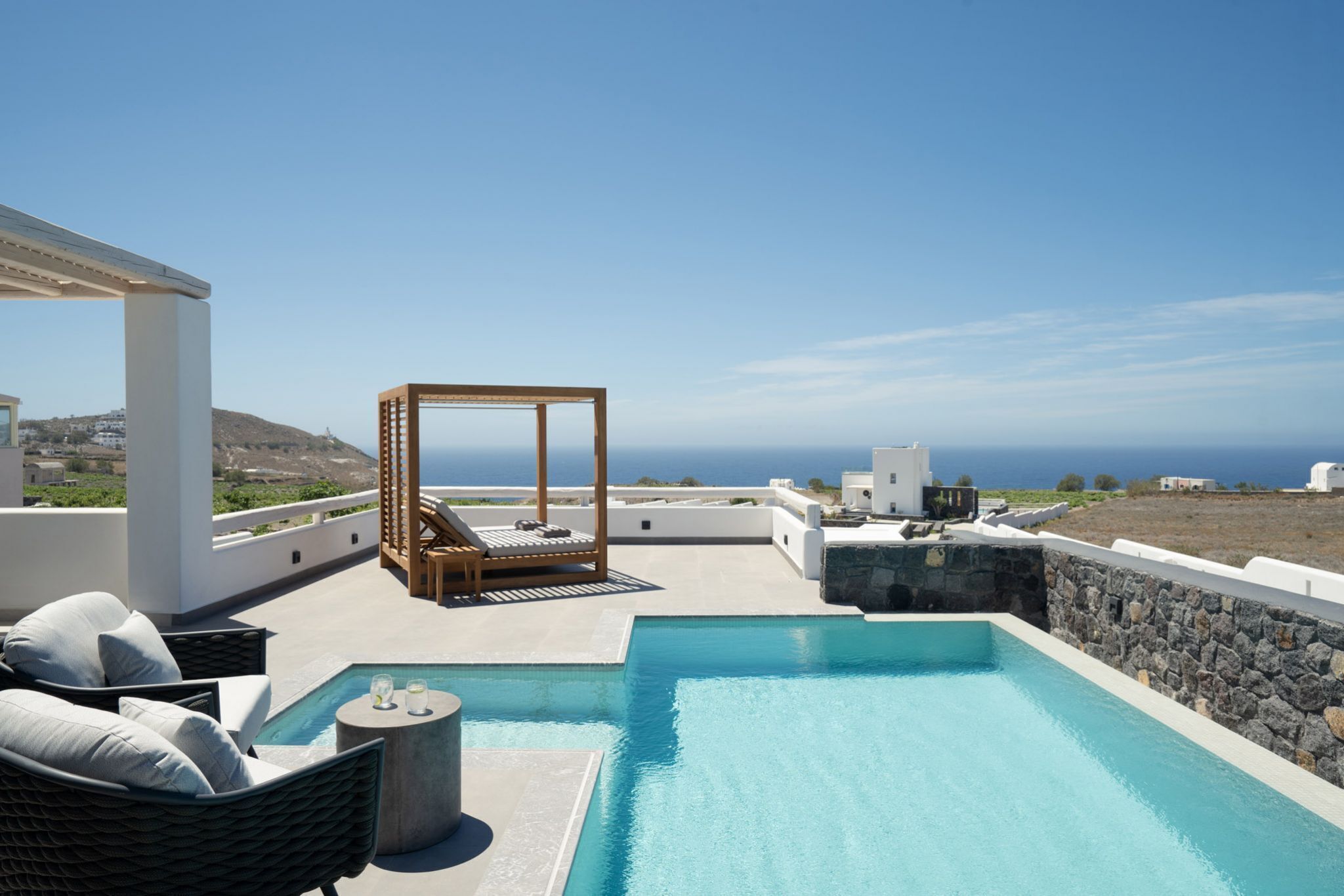 A rooftop pool area with a clear view of the ocean, a wooden lounge chair, and a stone wall.