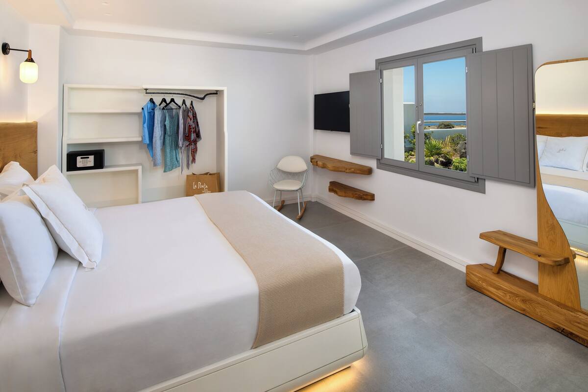 A modern bedroom with a large bed, a TV mounted on the wall, a window with a view of the sea, and a closet with hanging clothes.