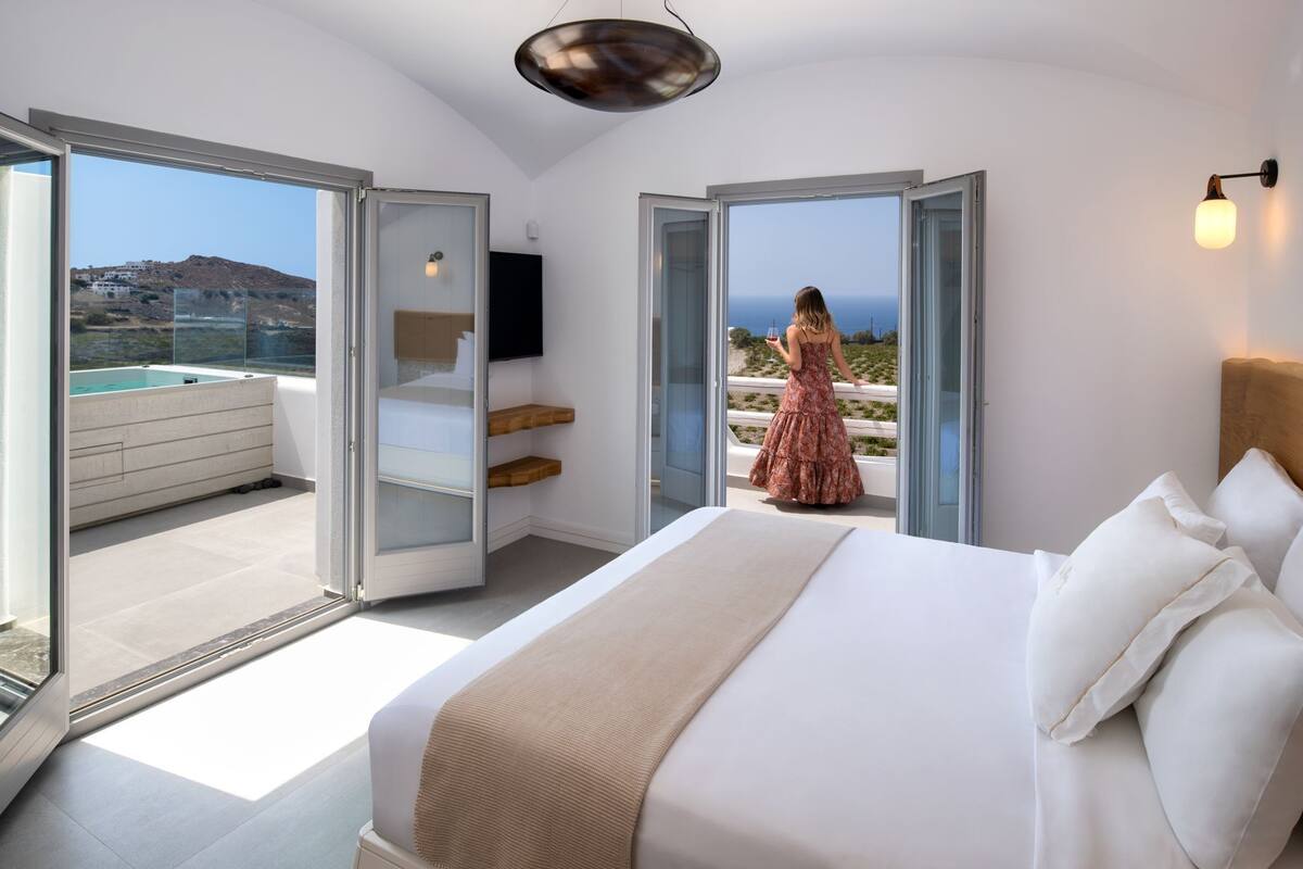 A bedroom with a large bed, a TV, and a balcony with a view of the sea.