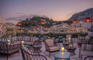 Un área de estar al aire libre con muebles de mimbre y lámparas rojas colgantes, con vista a un pueblo en la ladera.