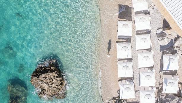 An aerial view of a beach with clear turquoise water, a rocky outcrop, and rows of white umbrellas arranged on the sand. - Hotel La Palma, Oetker Hotels (Capri)