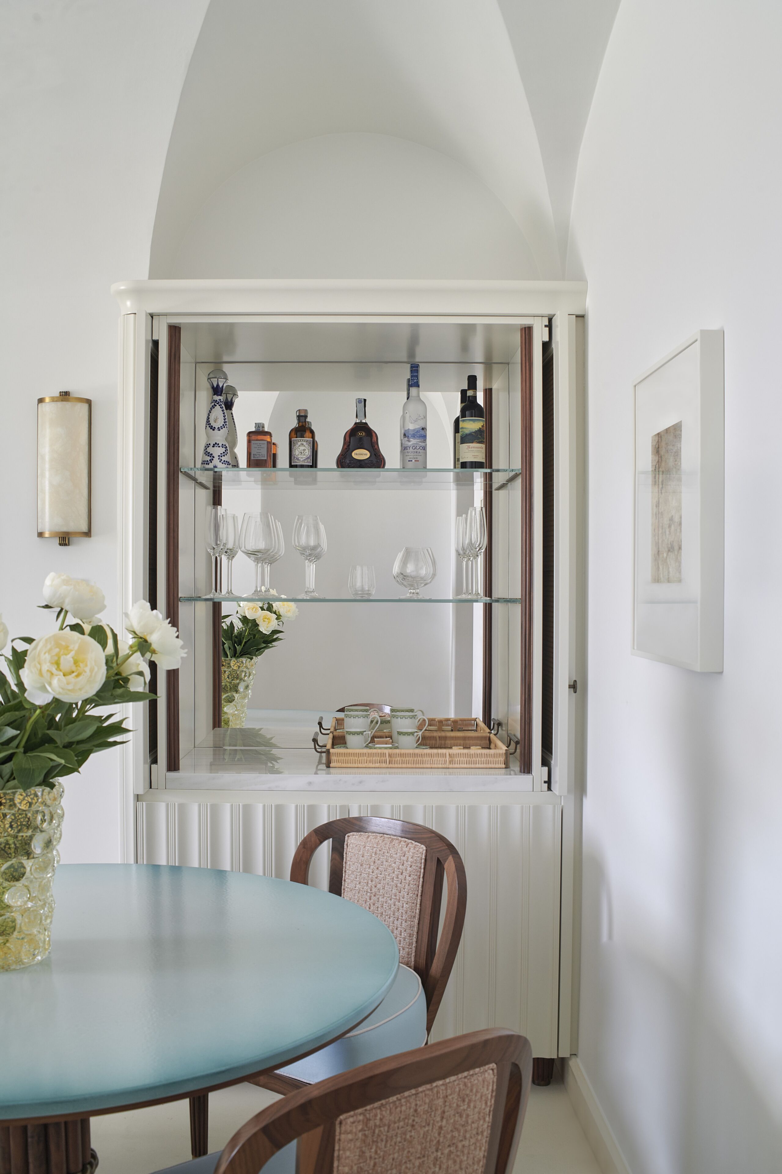 A dining area with a round table, chairs, a glass cabinet with bottles and glasses, and a vase with flowers.