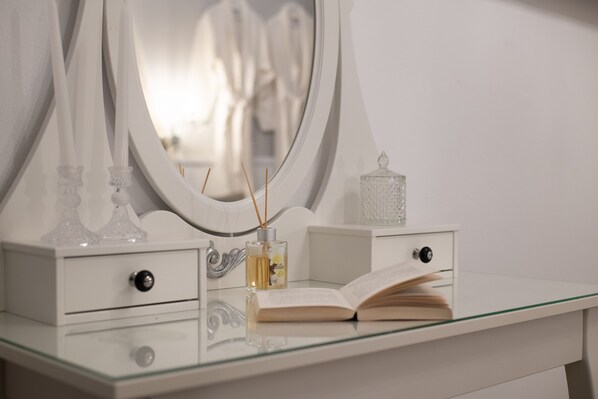 A white vanity with a glass top, a mirror, a book, a perfume bottle, and two candle holders. - Marikas Deluxe Rooms (Symi)
