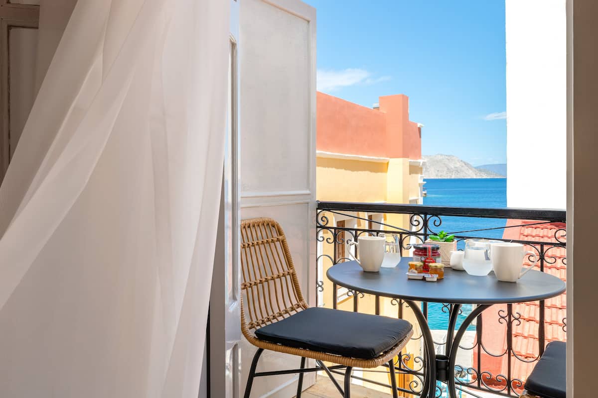 A balcony with a wicker chair and a small table, overlooking a coastal view with buildings and the sea.