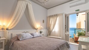 A bedroom with a large bed, a bedside table, a nightstand, and a view of the sea through an open door. - Marikas Deluxe Rooms (Symi)
