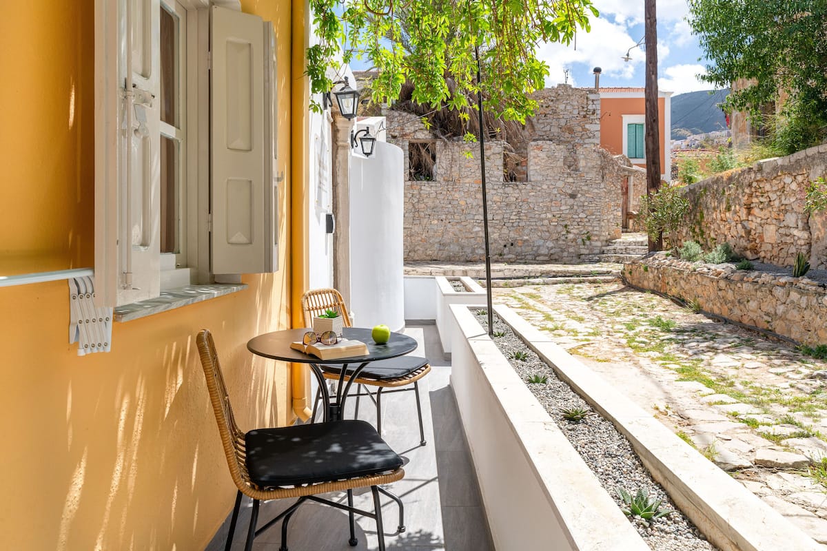 A small outdoor seating area with wicker chairs and a table, a stone wall, and a building with a yellow facade in the background.