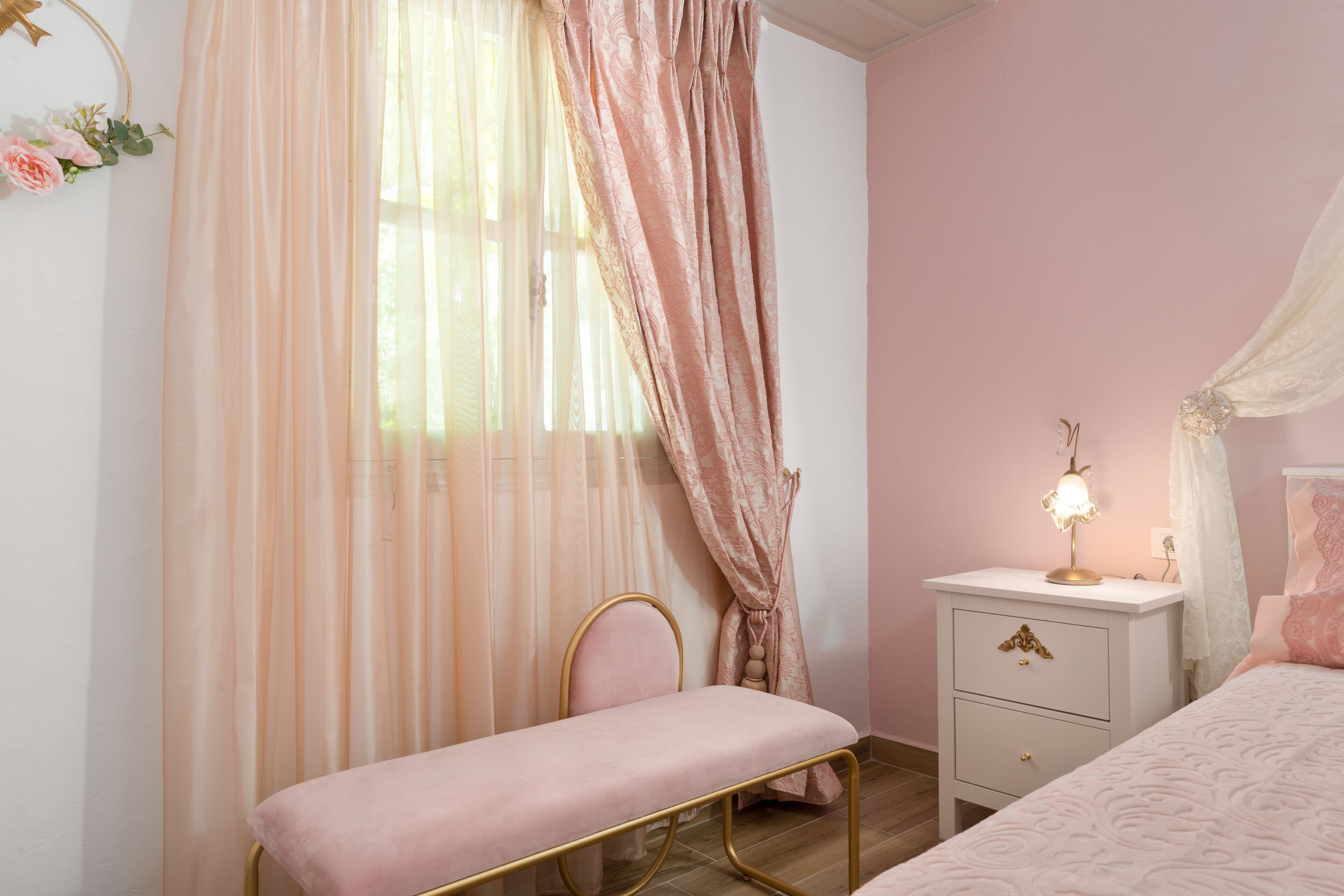 Une chambre à coucher décorée dans des tons roses, comprenant un lit, un banc, une table de chevet avec une lampe et des rideaux légers.