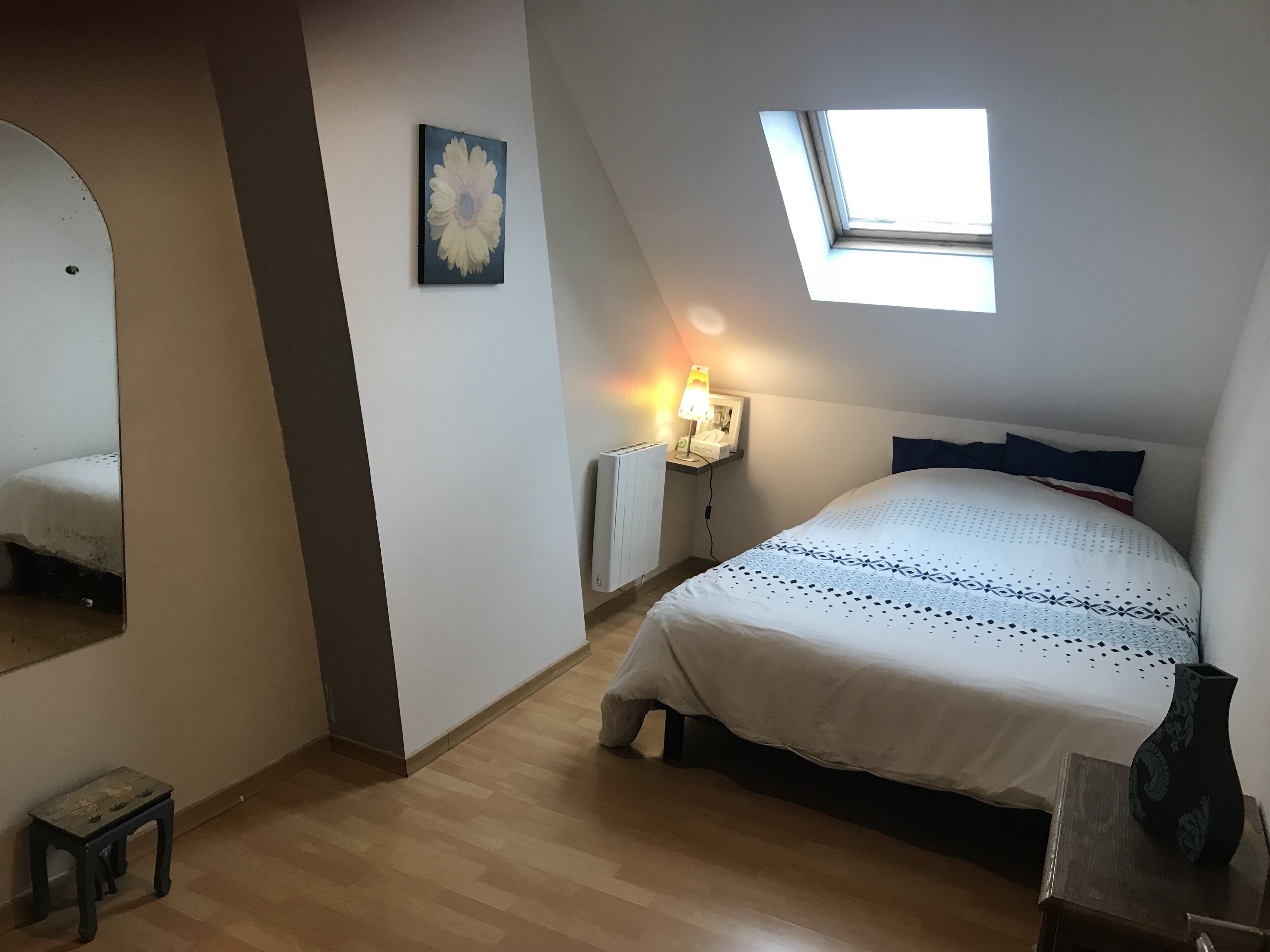 A bedroom with a sloped ceiling, a skylight, a bedside lamp, a bed with patterned bedding, a small table, and a mirror.