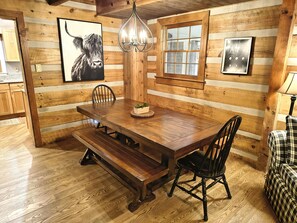 A wooden dining area with a bench, two chairs, a chandelier, and a window with blinds.