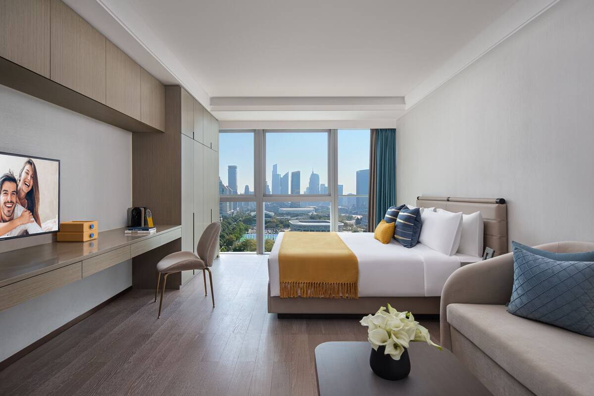 A modern hotel room with a large bed, a desk, and a view of the city skyline.