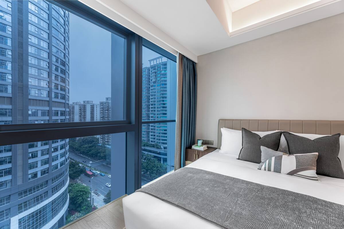 A modern hotel room with a large window, a bed with multiple pillows, a nightstand, and a view of the cityscape.