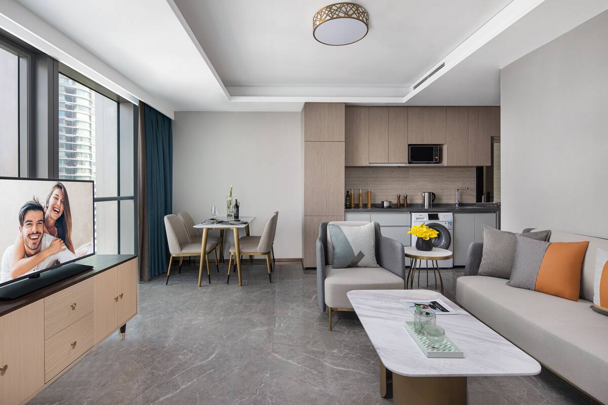 A modern living room with a sofa, a coffee table, and a TV displaying a couple. There is a dining area with chairs and a table, and a kitchen area with cabinets and appliances.