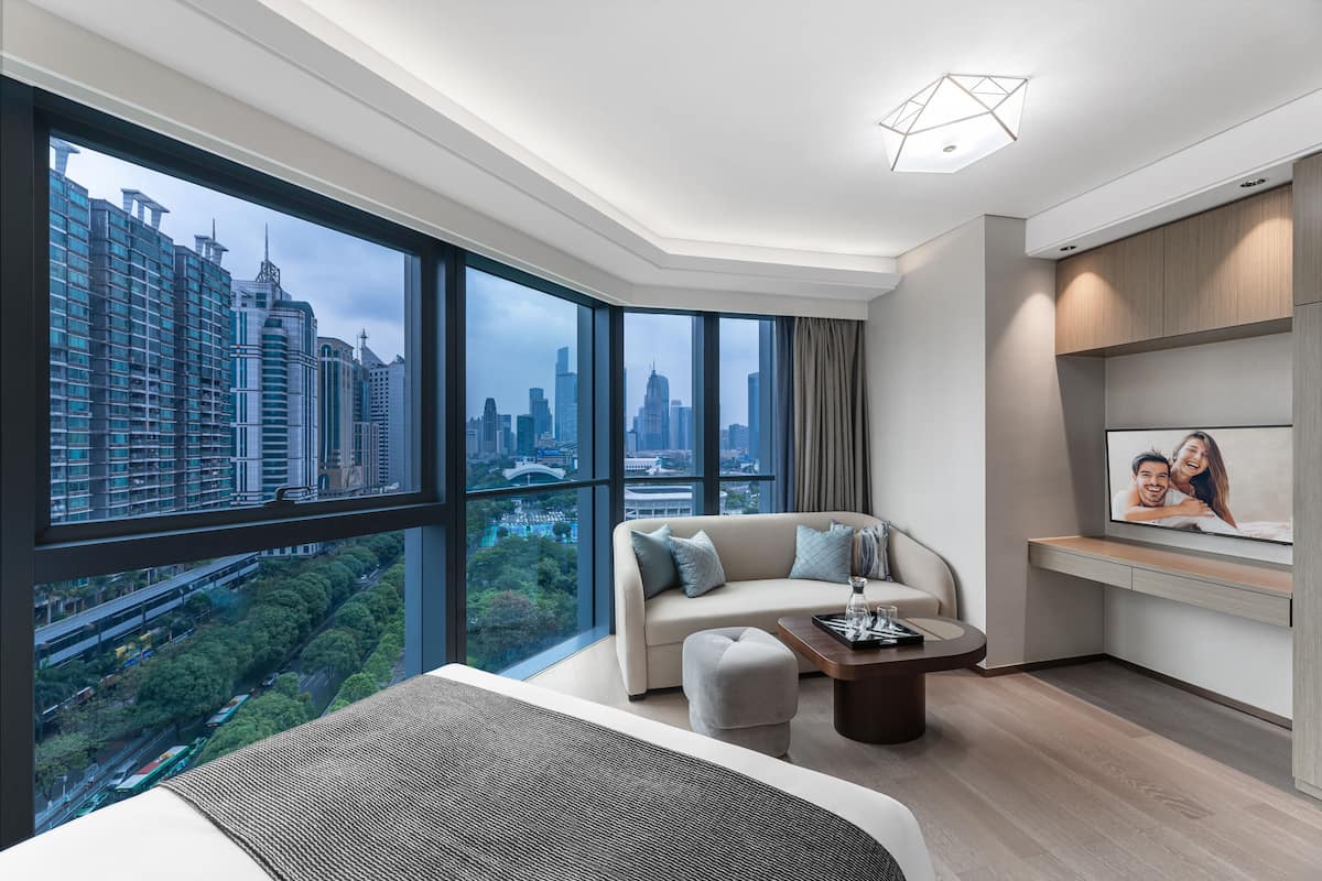 A modern hotel room with a large window offering a city view, a sofa, a coffee table, and a built-in TV.