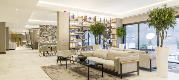 Un moderno vestíbulo de hotel con un sofá, sillas, una mesa de centro, una estantería y grandes ventanales.