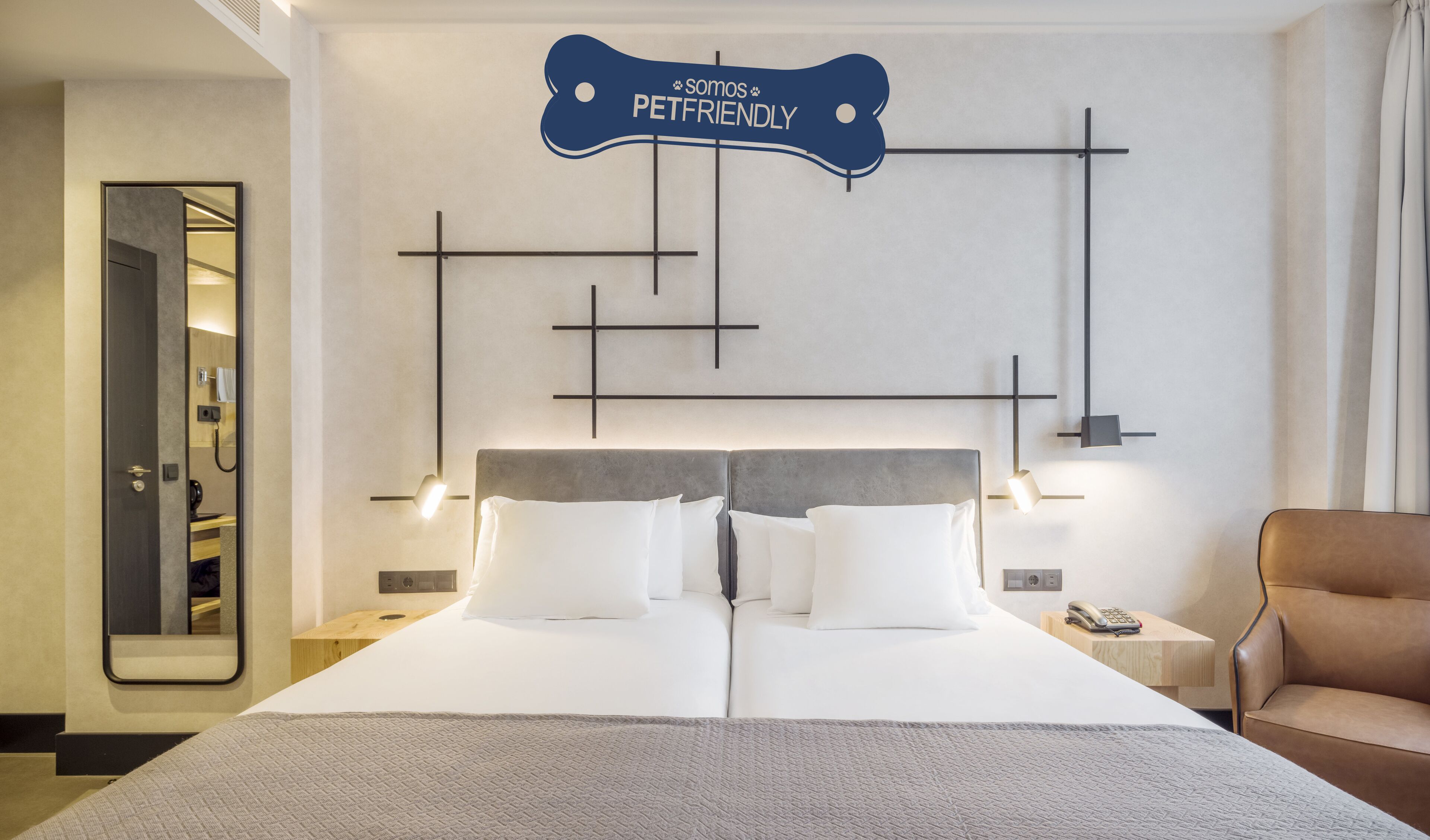 Habitación de hotel con diseño moderno, que incluye una cama grande con ropa blanca, cabecera gris y un letrero en la pared que dice “Somos PetFriendly”. Hay dos mesitas de noche con lámparas, un aparador de madera y un sillón de cuero.