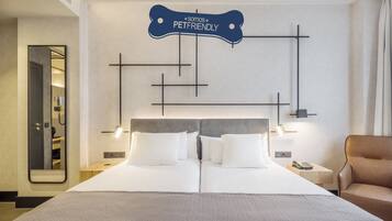 Habitación de hotel con diseño moderno, que incluye una cama grande con ropa blanca, cabecera gris y un letrero en la pared que dice “Somos PetFriendly”. Hay dos mesitas de noche con lámparas, un aparador de madera y un sillón de cuero.