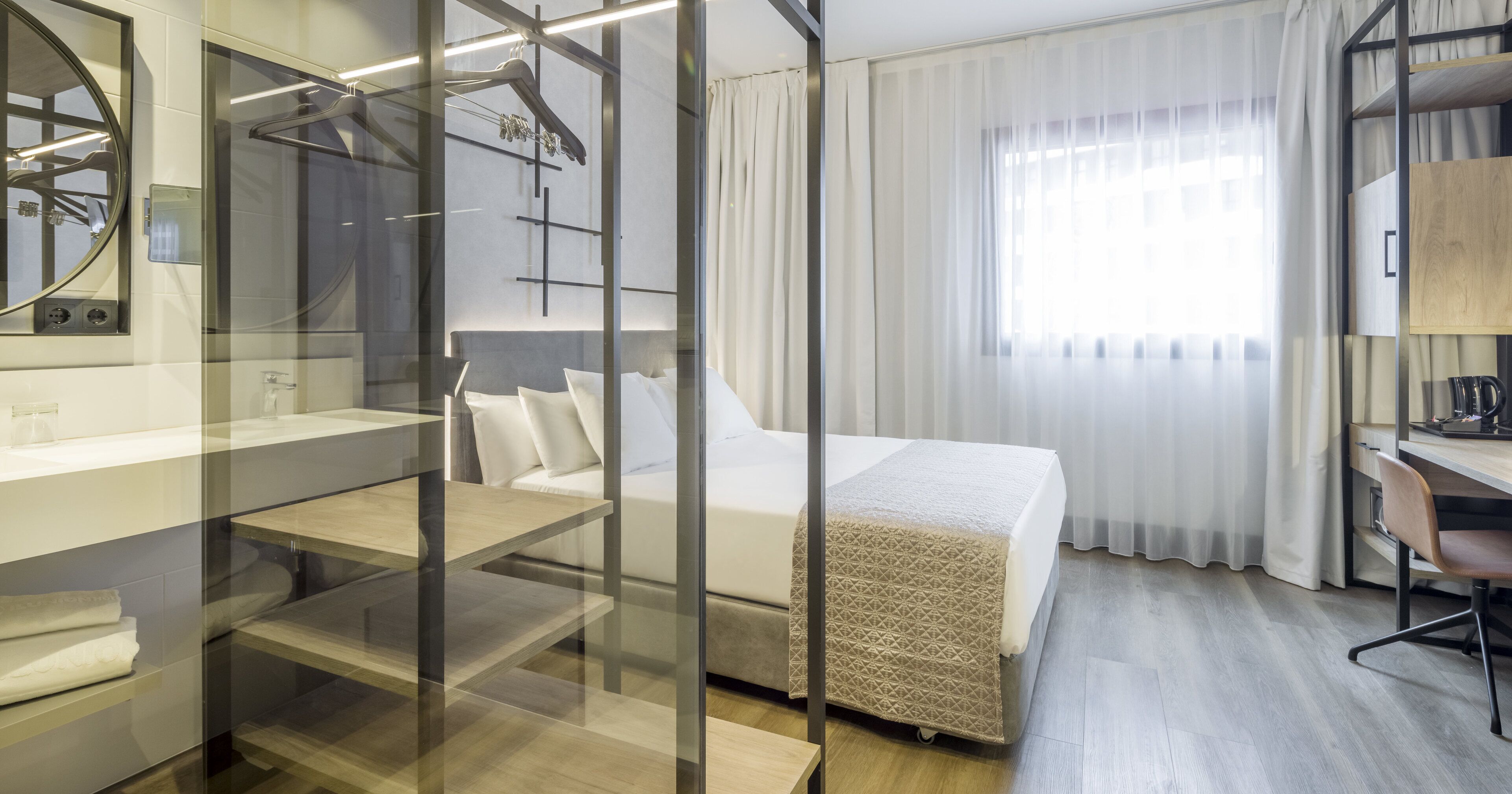 A modern hotel room with a bed, a desk, a chair, a mirror, and a shower area.