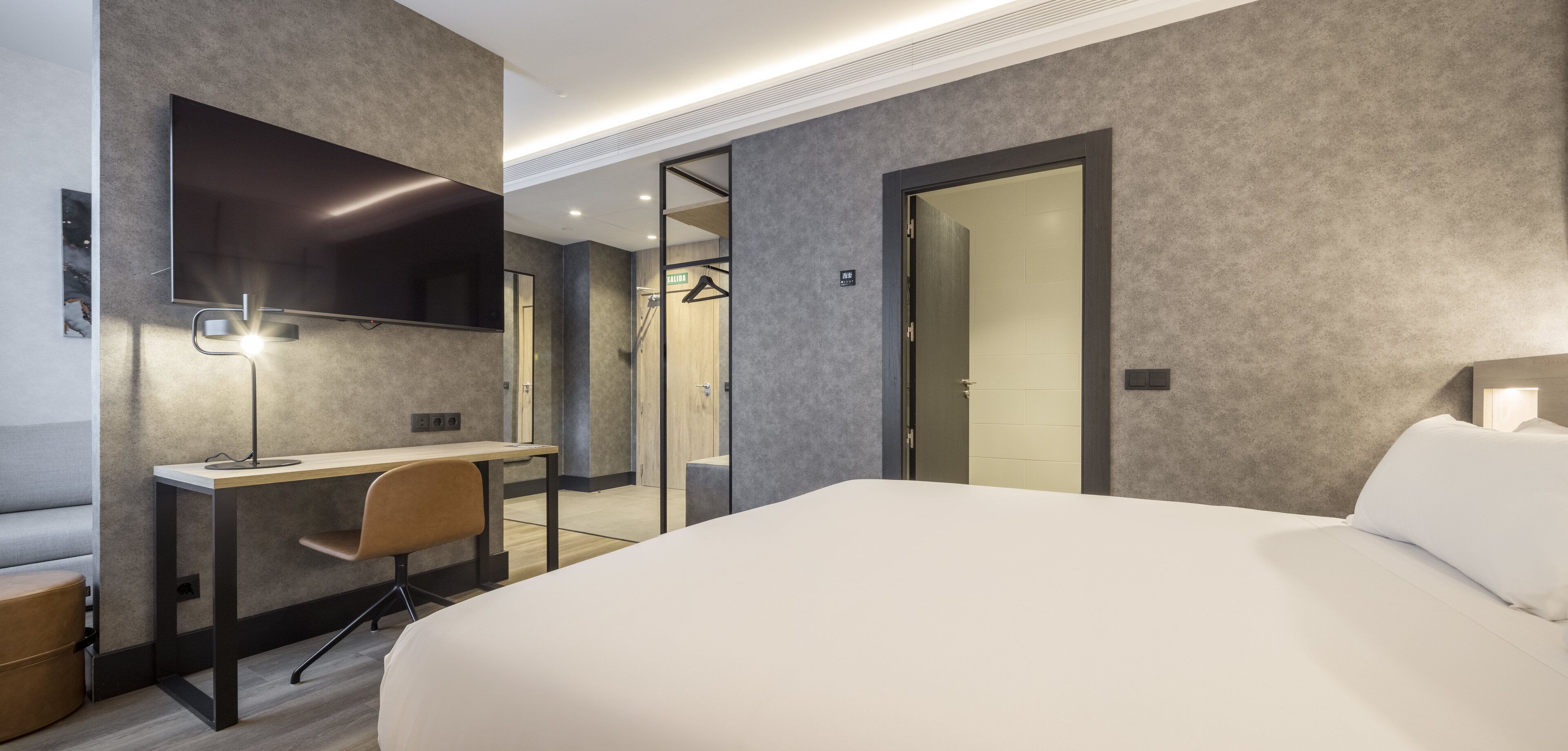 Una habitación de hotel moderna con una cama grande, un escritorio con silla, un televisor de pantalla plana en la pared y un vestidor.
