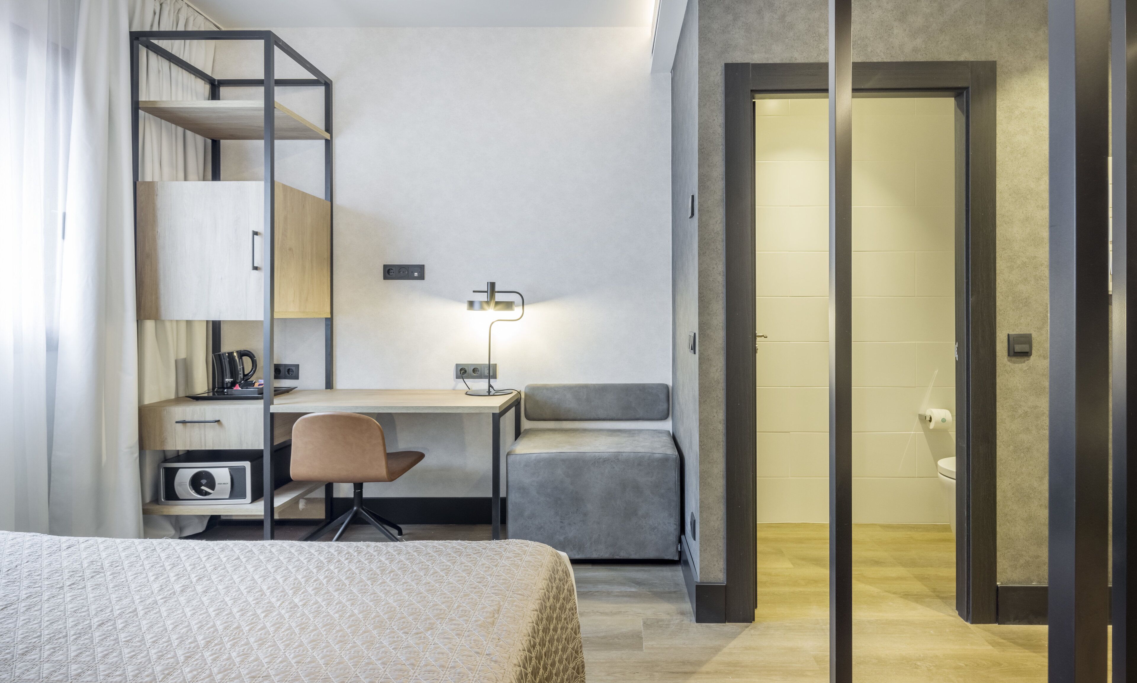 Habitación de hotel moderna con una cama, un escritorio, una silla, un armario y un baño visible a través de una puerta abierta.