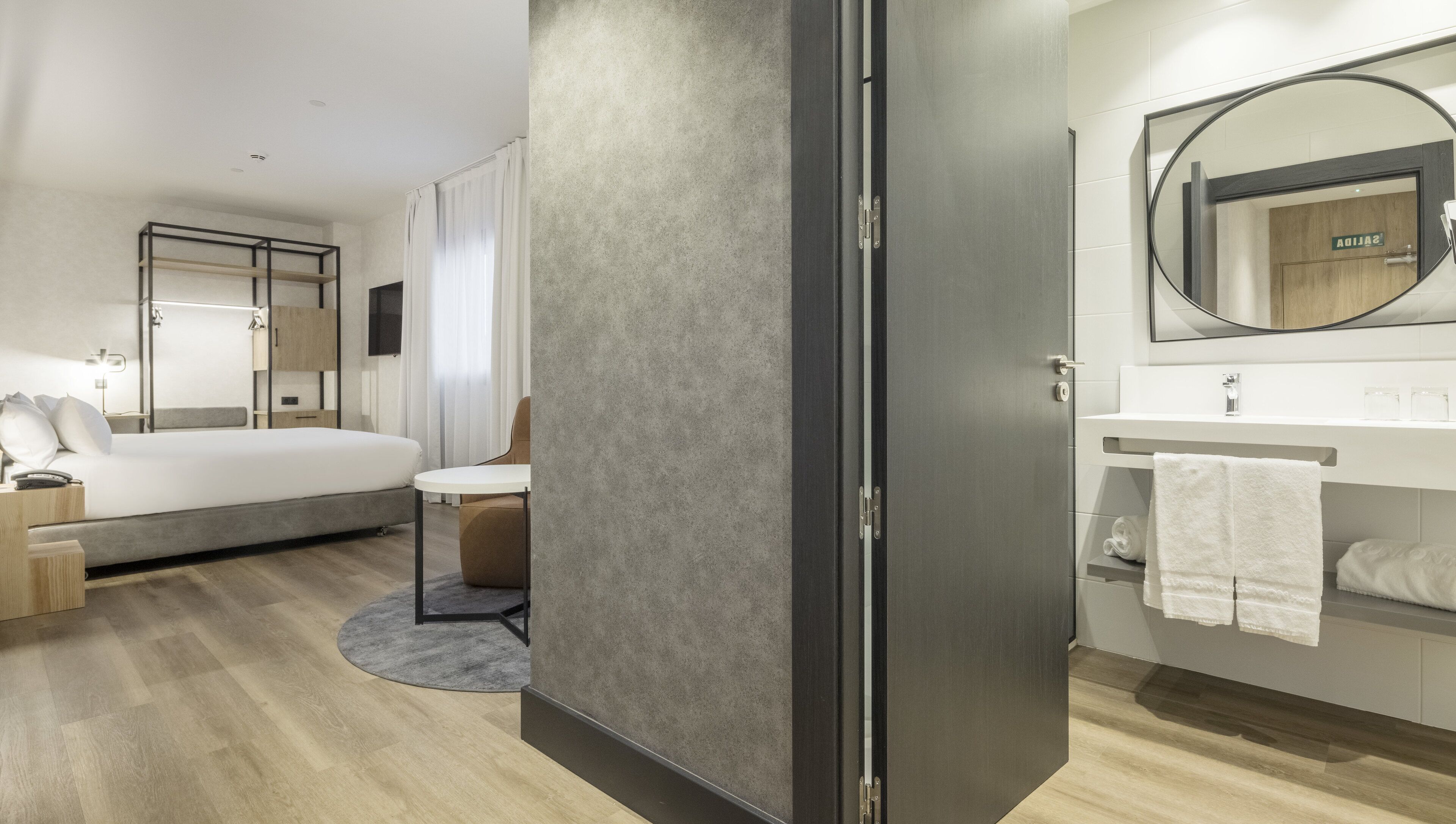 A modern hotel room with a bed, a desk, a chair, a round mirror, and a bathroom with a sink and towel rack.