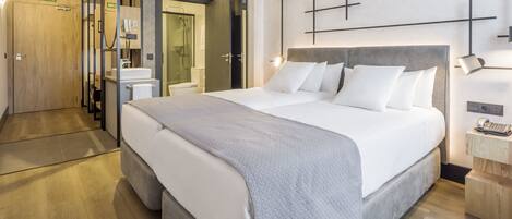 A modern hotel room with a large bed, a bedside table with a lamp, and a wooden wardrobe.