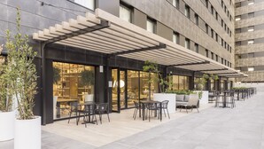 Un área de café al aire libre con mesas y sillas, una zona cubierta para sentarse y plantas en macetas.