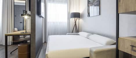 A modern hotel room with a large bed, a desk with a chair, a standing lamp, and a flat-screen TV mounted on the wall.