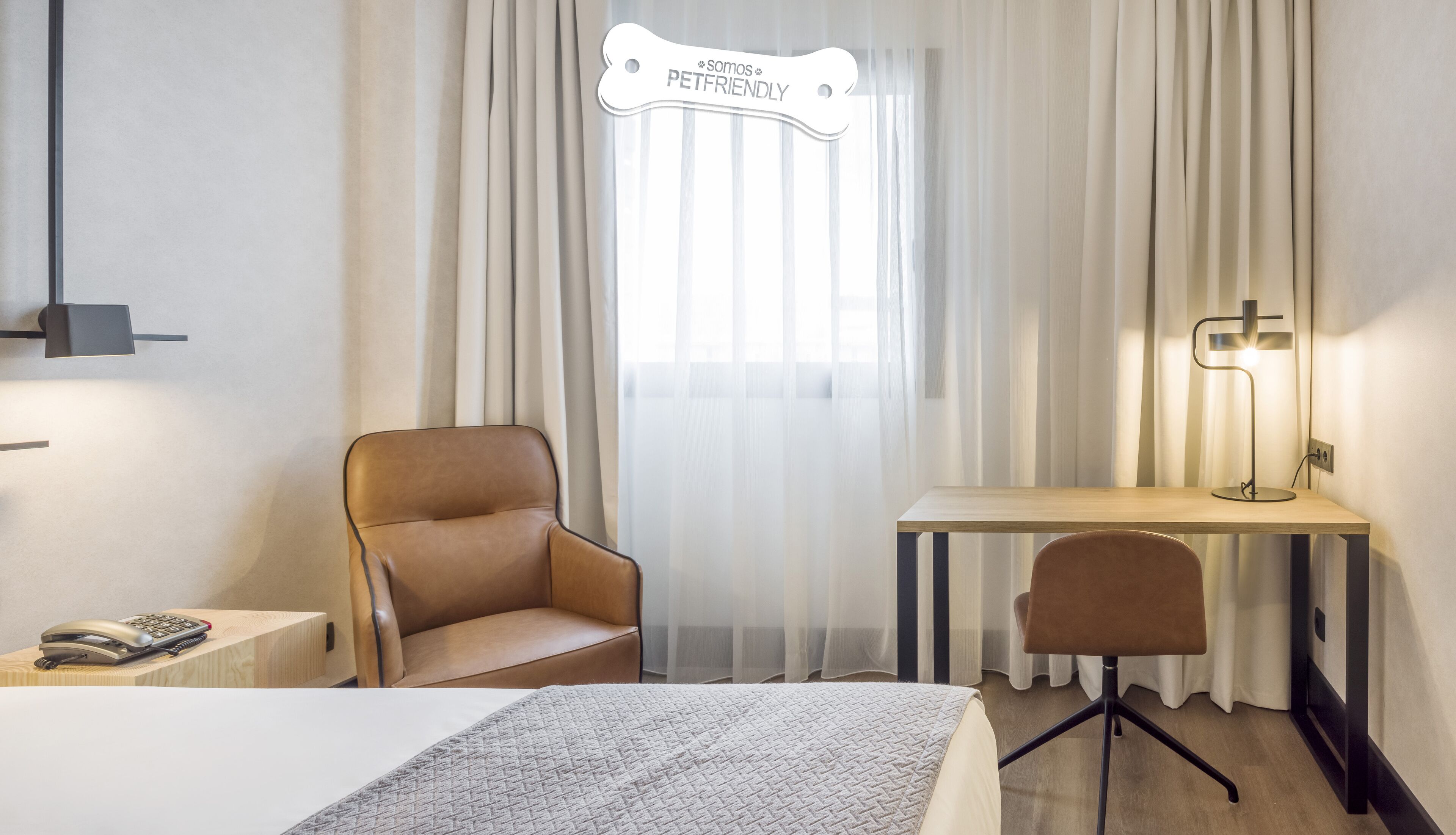Habitación de hotel con cama, escritorio, silla, teléfono en la mesita de noche y un cartel sobre la ventana que dice "Somos PETFRIENDLY".