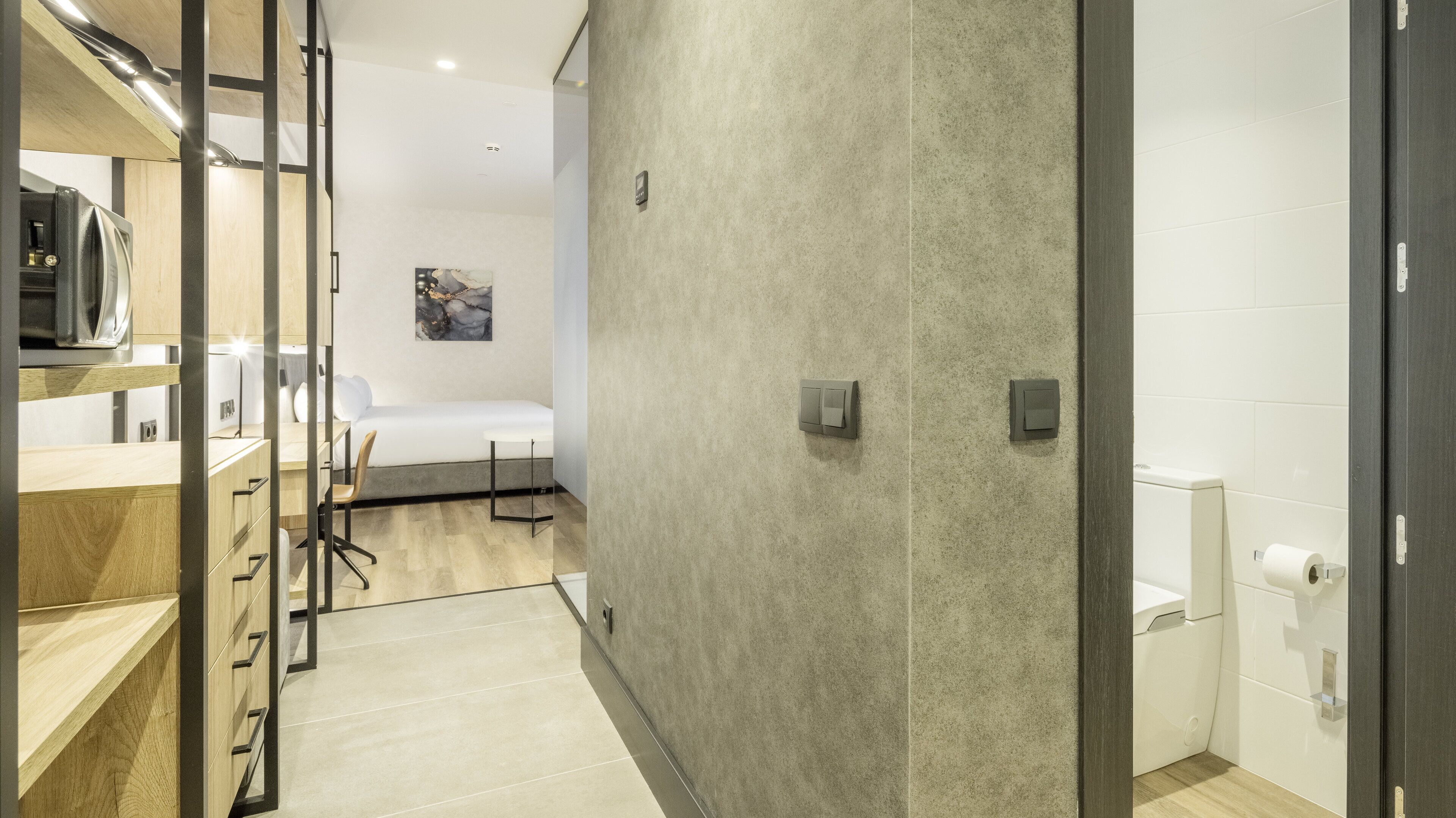 Una habitación de hotel moderna con kitchenette, una cama, una mesita y un baño con inodoro y lavamanos.