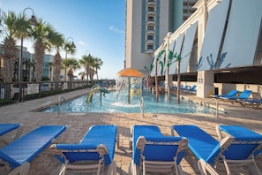 Zona de piscina con sillones azules, fuente de agua, palmeras y un edificio con balcones.