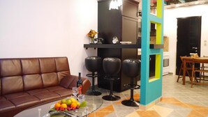 A modern living room with a brown leather sofa, a glass coffee table with fruit, black bar stools, and a teal accent wall.