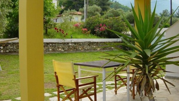 A covered patio with yellow chairs and a table, overlooking a garden with a mountain in the background.