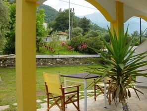 Un porche cubierto con sillas amarillas y una mesa, con vistas a un jardín y una montaña al fondo.