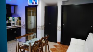 A modern living room with a glass dining table, wooden chairs, a white sofa, and a TV mounted on the wall.