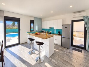 A modern kitchen with a central island, bar stools, a built-in oven, and a view of a swimming pool. - Villa Serenity – Stylish 3-Bedroom Retreat with Private Pool (Vodnjan)