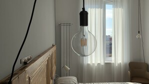 Ein Schlafzimmer mit Holz-Kopfteil, einem Bett mit gestreifter Bettwäsche, einer Pendelleuchte, einem Heizkörper und Blick ins Freie durch durchsichtige Vorhänge.