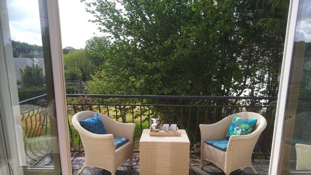 Un balcon avec des fauteuils en osier et une petite table, un sol en pierre et une vue sur la verdure et les bâtiments en arrière-plan.