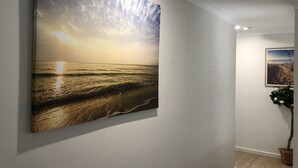 En hall med en stor tavla som föreställer ett strandlandskap i solnedgång, en mindre inramad bild på väggen och en krukväxt.