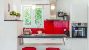 A modern kitchen with a central island, two red bar stools, a window with a view of greenery, and a built-in oven. - Villa Sofia (Santalezi)