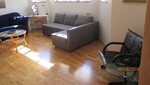 Ein Wohnzimmer mit grauem Sofa, blauem Sessel, einem Glastisch und einem schwarzen Bürostuhl. Zwei Fenster mit Jalousien, eine Heizung und ein Bild an der Wand sind ebenfalls zu sehen.