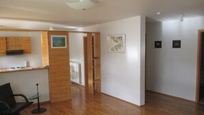 Ein geräumiges Zimmer mit Holzboden, einer Küchenzeile mit Holzausstattung und einem Kühlschrank. An der Wand befinden sich zwei gerahmte Kunstwerke und eine Tür, die in einen anderen Raum führt.