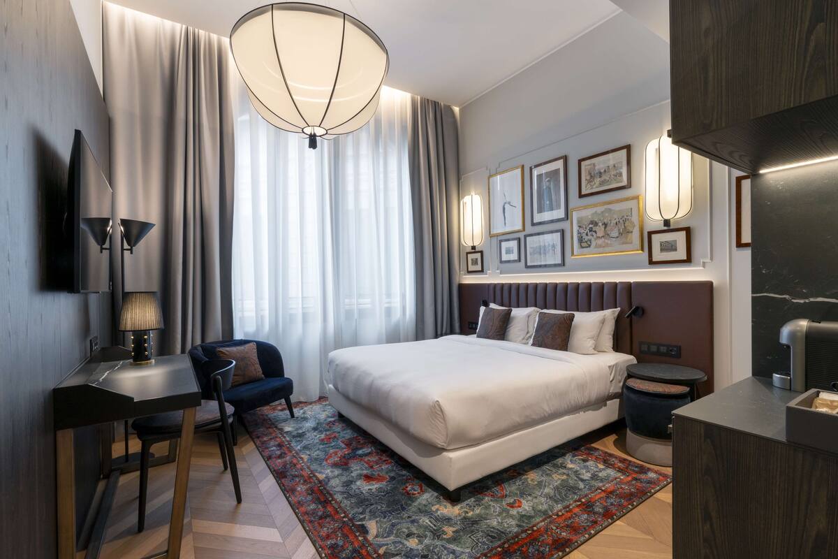 A modern hotel room with a large bed, a desk, a chair, a desk lamp, a TV, a bedside table, a nightstand, a rug, and framed pictures on the wall.
