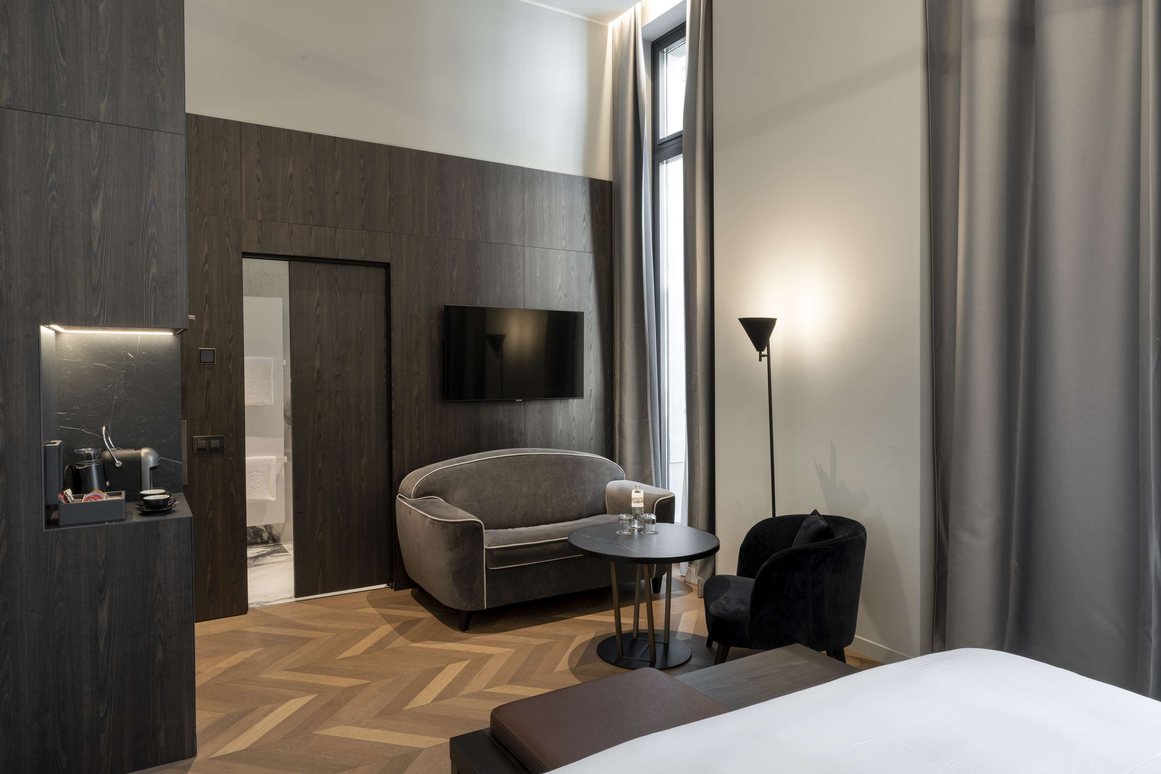 Une chambre d’hôtel moderne avec un grand lit, un canapé, une petite table, un téléviseur fixé au mur et une fenêtre avec des rideaux.