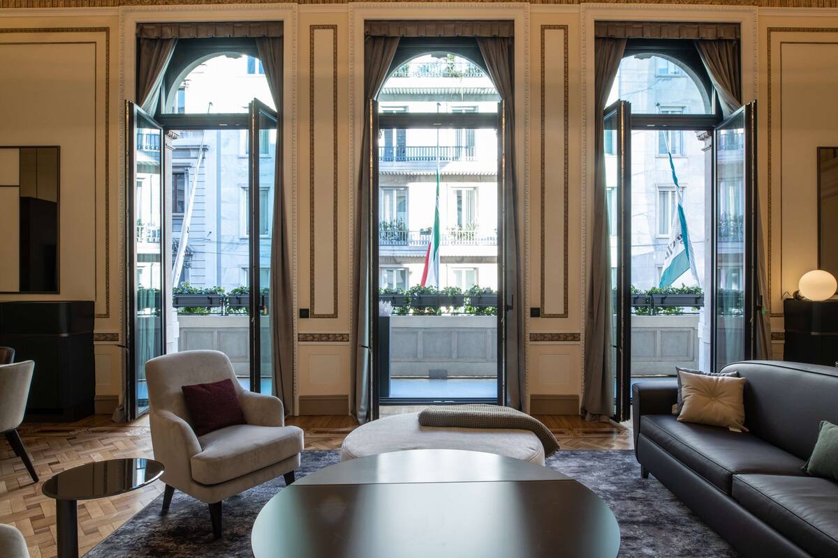 A spacious living room with a sofa, armchair, and ottoman, large windows with curtains, and a view of the cityscape.
